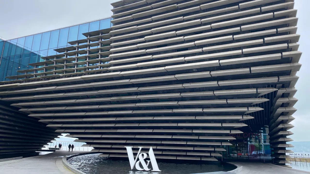 Exterior of V&A Dundee museum showing the layered, angular façade above the entrance, with the ‘V&A’ sign beside a reflecting pool.