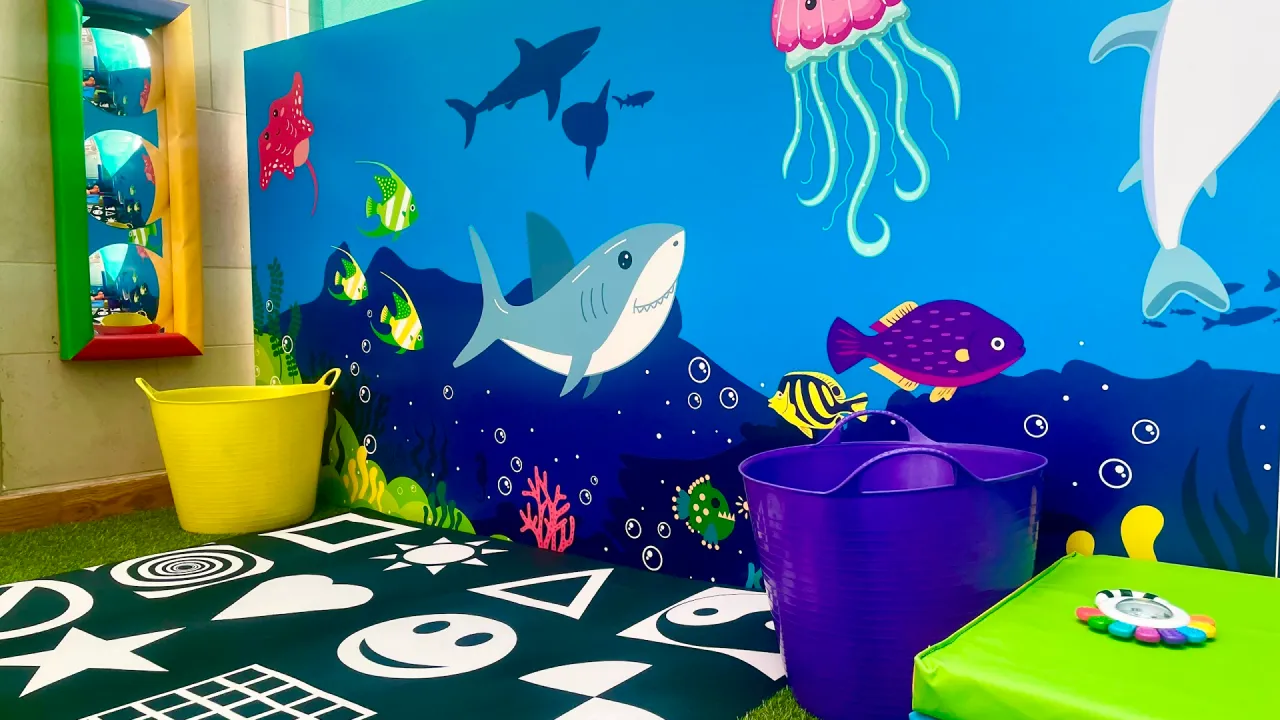 Underwater sea-life mural with a shark, fish, and jellyfish above colourful buckets in the children’s play area at Dundee Olympia Leisure Pool.