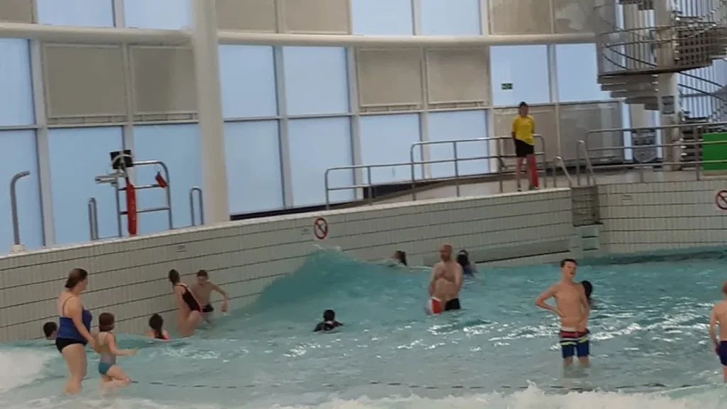 Swimmers enjoying the wave pool at Dundee Olympia Leisure Pool, with a visible wave cresting near the wall and a lifeguard standing on the poolside platform.