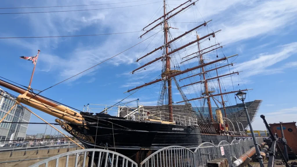 RRS Discovery tall ship moored at Dundee waterfront, with its masts and rigging against a blue sky, and ‘DISCOVERY’ visible on the hull.