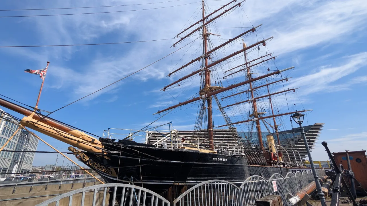 RRS Discovery tall ship moored at Dundee waterfront, with its masts and rigging against a blue sky, and ‘DISCOVERY’ visible on the hull.