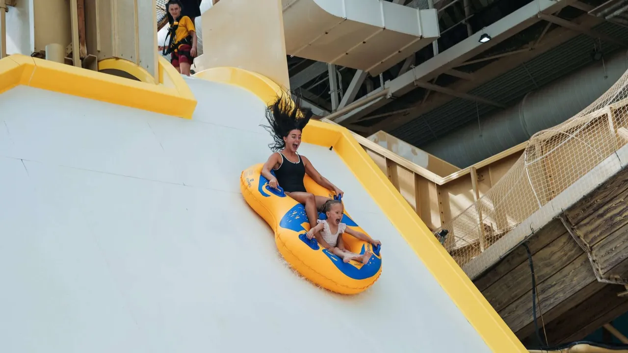 Adult and child riding a two-person inflatable ring down a steep white flume slide at Sandcastle Waterpark in Blackpool, with a lifeguard at the top platform.
