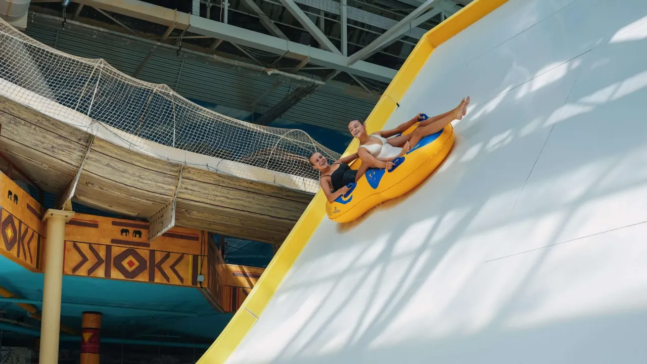 Two riders on a yellow two-person inflatable ring descending a steep white flume slide inside Sandcastle Waterpark, Blackpool.