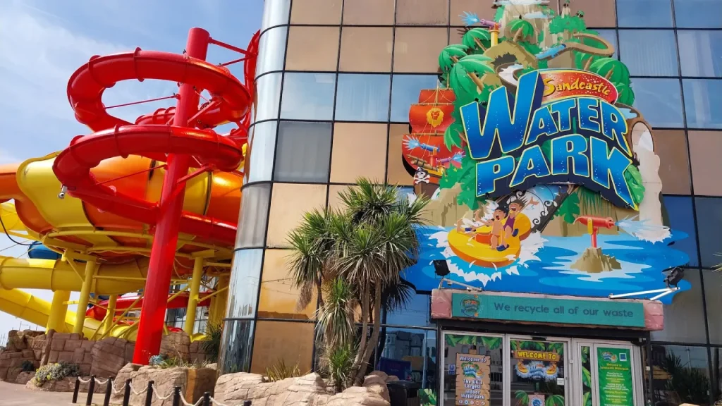Front entrance of Sandcastle Waterpark in Blackpool with the large ‘Sandcastle Water Park’ sign on the building and red, yellow, and orange outdoor flume slides beside it.
