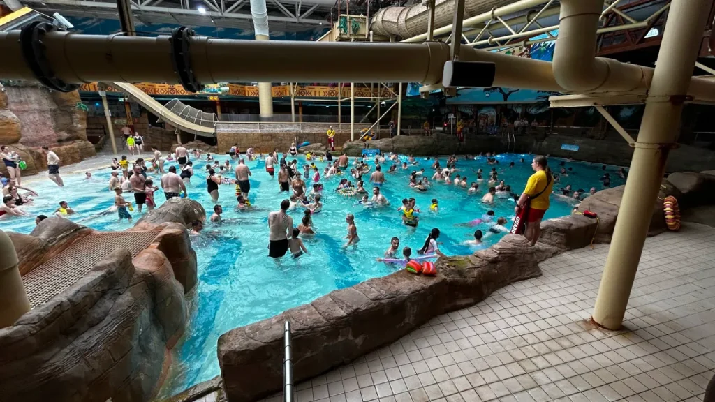 Crowded indoor pool at Sandcastle Waterpark in Blackpool with swimmers across the water and a lifeguard standing on the poolside near rock-effect edging.