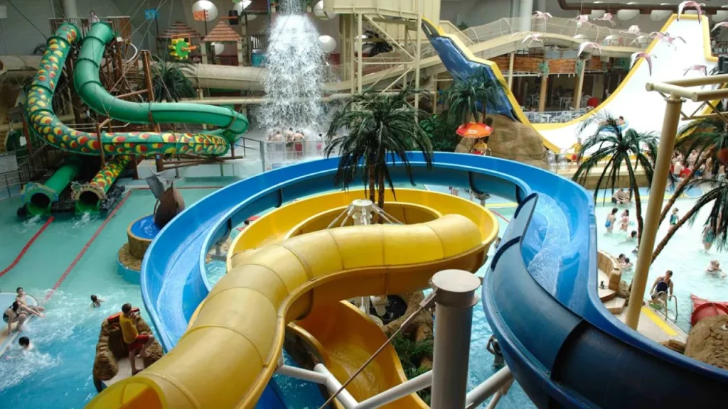 Close-up view of blue and yellow twisting flume slides inside Sandcastle Waterpark in Blackpool, with a large water feature, palm trees, and swimmers in the pools below.