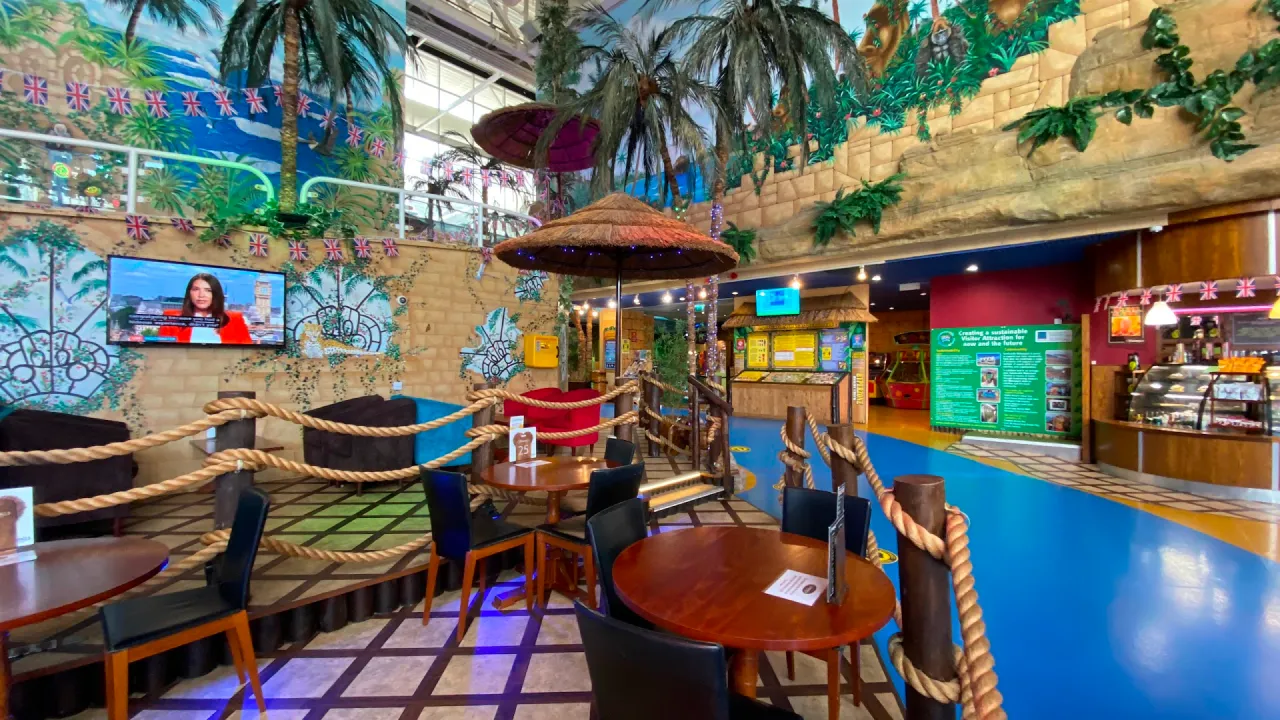 Tropical-themed café seating area inside Sandcastle Waterpark in Blackpool, with palm-tree décor, rope railings, round tables, wall murals, and the food counter in the background.