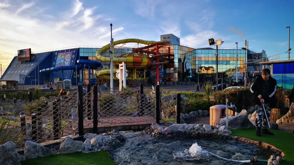 Wide exterior view of Sandcastle Waterpark in Blackpool showing the glass-fronted building and outdoor yellow and red flume slides, with a small pond and rope bridge in the foreground.