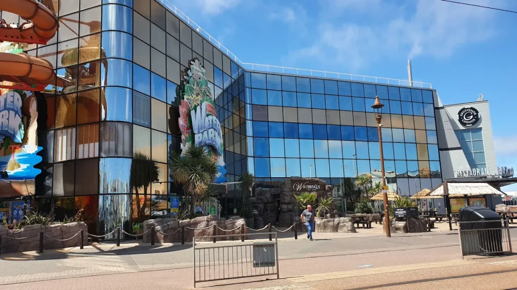 Exterior of Sandcastle Waterpark in Blackpool showing the reflective glass building, large ‘Water Park’ sign, rock-style Waterfalls Café-Bar entrance, and palm trees on the promenade.