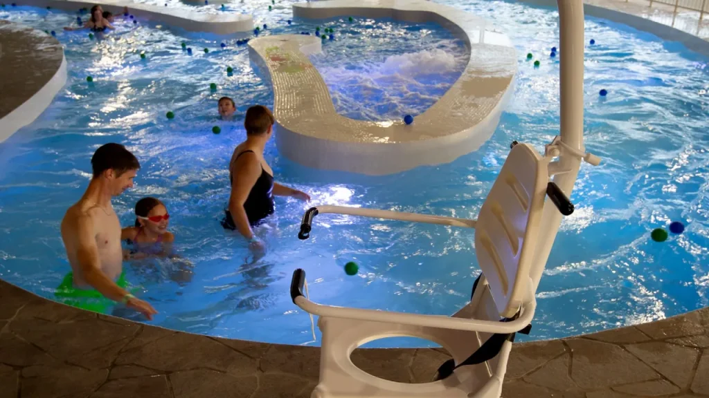 Pool hoist chair beside the indoor pool at The Wave Waterpark in Coventry, with adults and children swimming in shallow water near an island feature.