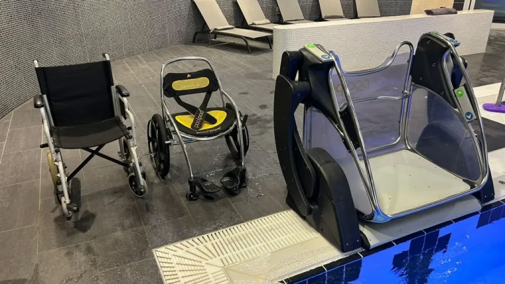 Pool pod transfer system beside the water at The Wave Waterpark in Coventry, with a wheelchair and a poolside shower chair on the wet deck nearby.