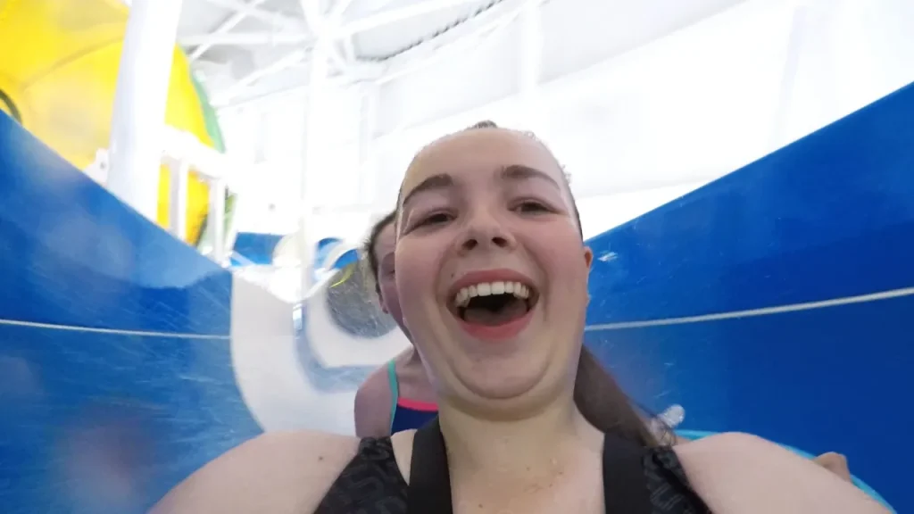 Smiling rider taking a selfie while travelling down an open blue flume slide at The Wave Waterpark in Coventry, with another rider behind.