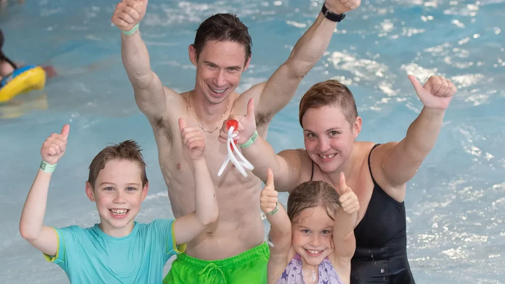 Smiling family of four in the pool at The Wave Waterpark in Coventry, raising their arms and giving thumbs up in the water.