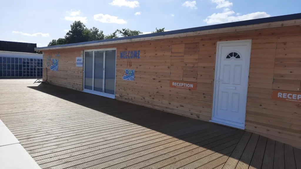 Wooden Aqua Action reception building at Fairlop Waters with ‘Welcome’ and ‘Reception’ signs, next to a wide decked walkway under a blue sky.