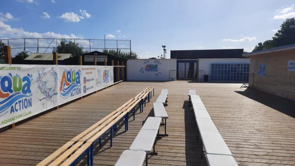 Aqua Action Fairlop Waters outdoor deck with long benches and tables, branded wall banners, and the main activity buildings in the background on a sunny day.