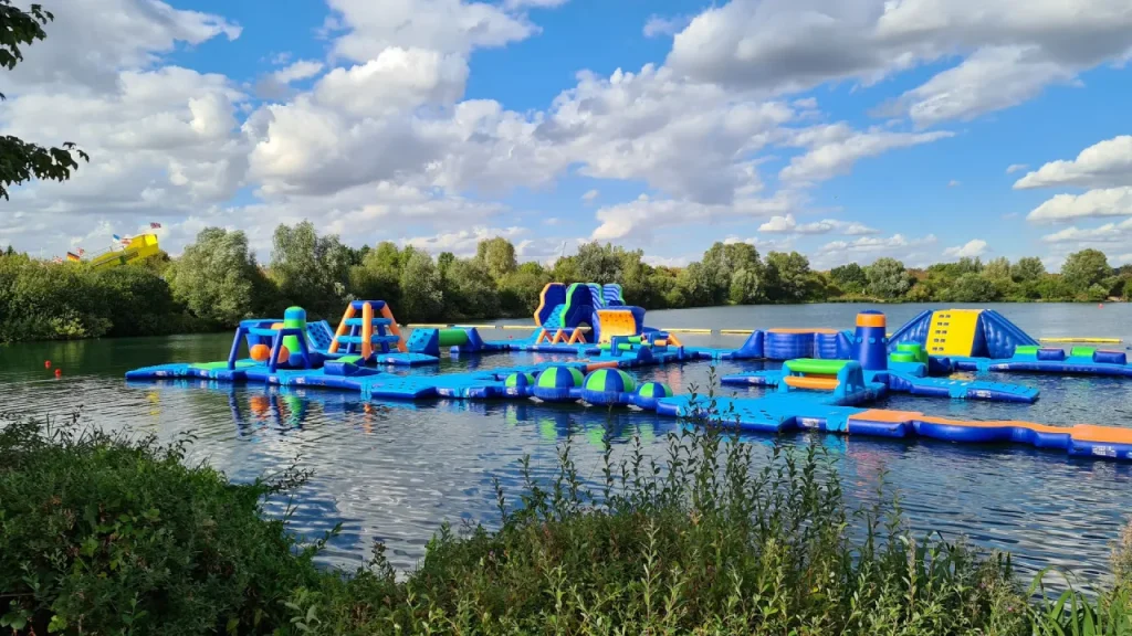Wide view of the Aqua Action Fairlop Waters inflatable aqua park on the lake, with blue, green, and orange obstacle sections, trees along the shoreline, and a partly cloudy sky.