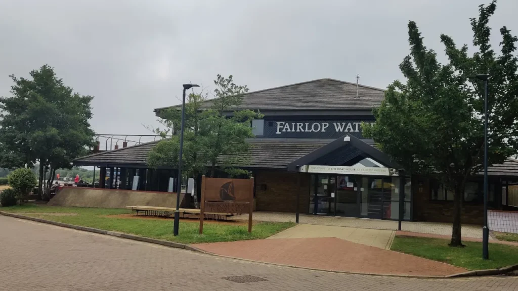 Exterior view of the Fairlop Waters Boathouse building and entrance under an overcast sky, with the Fairlop Waters sign, trees, and a walkway in the foreground.