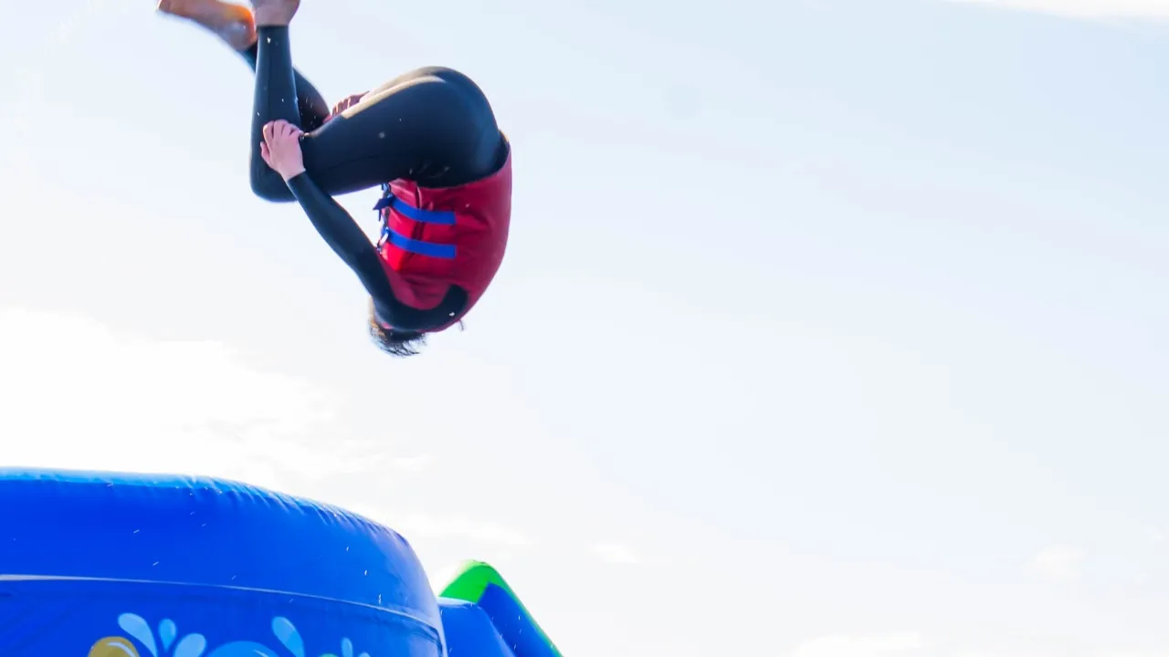 Participant in a red buoyancy aid and black wetsuit mid-air in a tucked flip above a blue inflatable obstacle at Aqua Action, Fairlop Waters, against a bright sky.