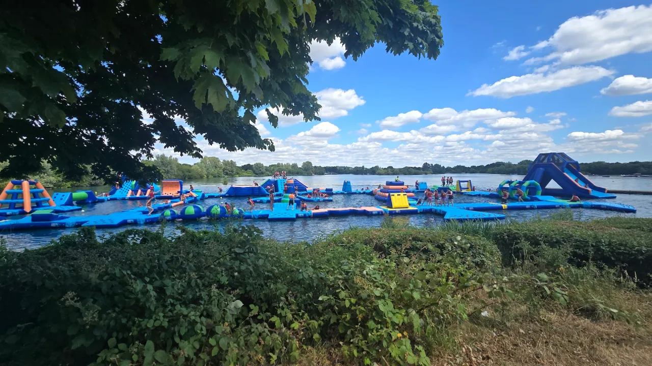 Wide shoreline view of the Aqua Action inflatable aqua park at Fairlop Waters, with blue floating obstacles, slides, and groups of visitors on the course under a bright sky.