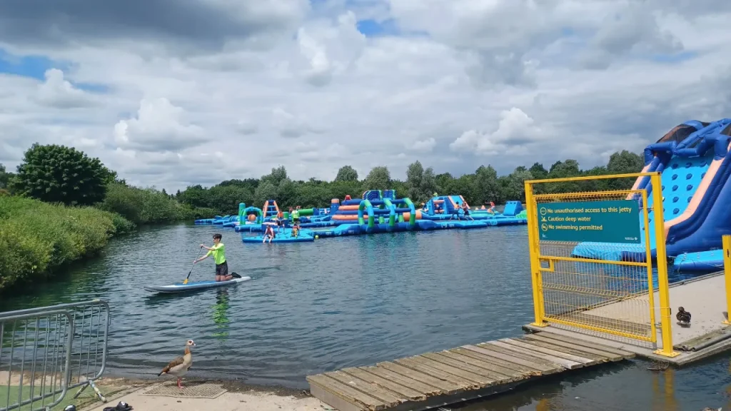 View across Fairlop Waters towards the Aqua Action inflatable aqua park, with blue floating obstacles and a large slide, a paddleboarder on the lake, and a yellow-gated jetty in the foreground.
