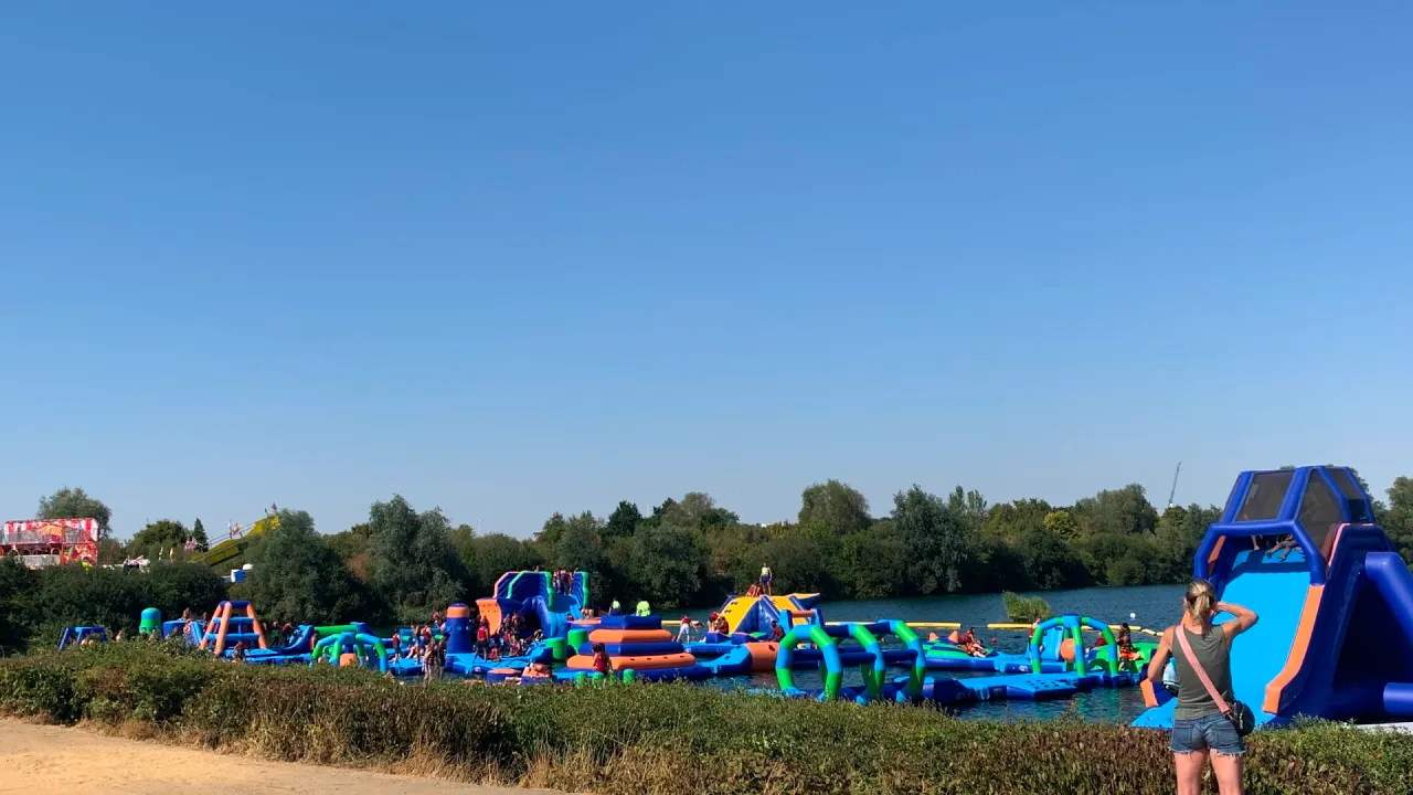 Wide view of the Aqua Action inflatable aqua park at Fairlop Waters, with blue, green, and orange floating obstacles and a tall slide on the lake, framed by the lakeside path and trees.