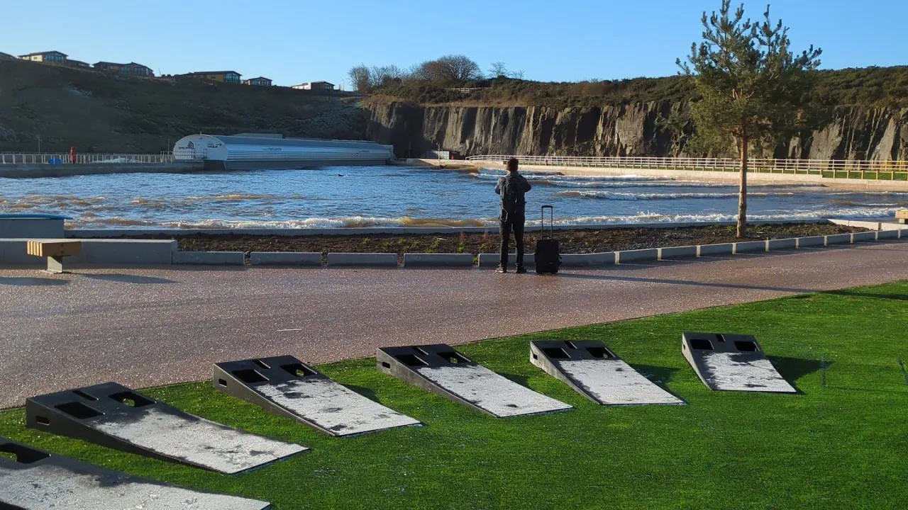 Wide view of the Lost Shore Surf Resort surf lagoon in Scotland, showing waves breaking across the artificial surf pool, rock wall scenery, a visitor watching from the promenade, and cornhole boards on the grass in the foreground.