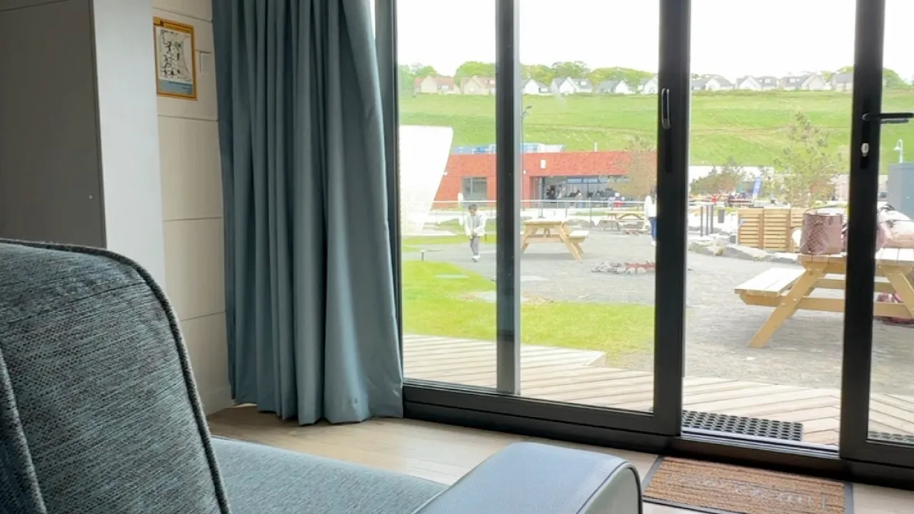 Interior view from Lost Shore Surf Resort accommodation in Scotland, showing a sofa beside curtains and large sliding glass doors looking out to the outdoor courtyard, picnic tables, and grassy hills with homes in the distance.