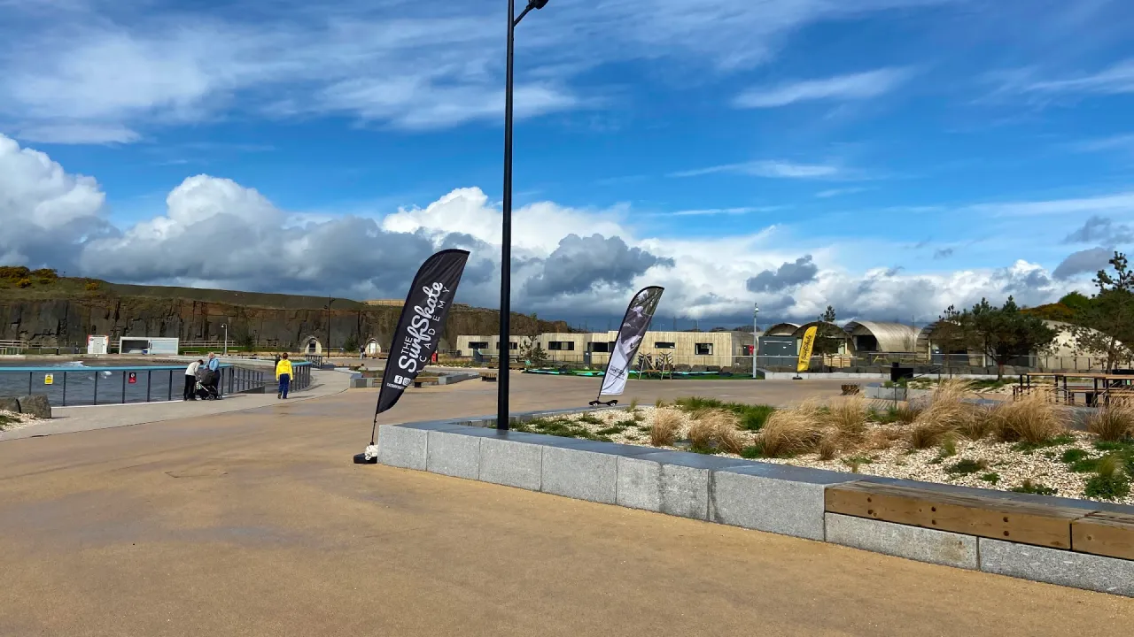 Wide paved promenade at Lost Shore Surf Resort in Scotland, with feather flags along the walkway, modern resort buildings in the background, landscaped planters and seating, and the surf lagoon and railings to the left under a bright, partly cloudy sky.