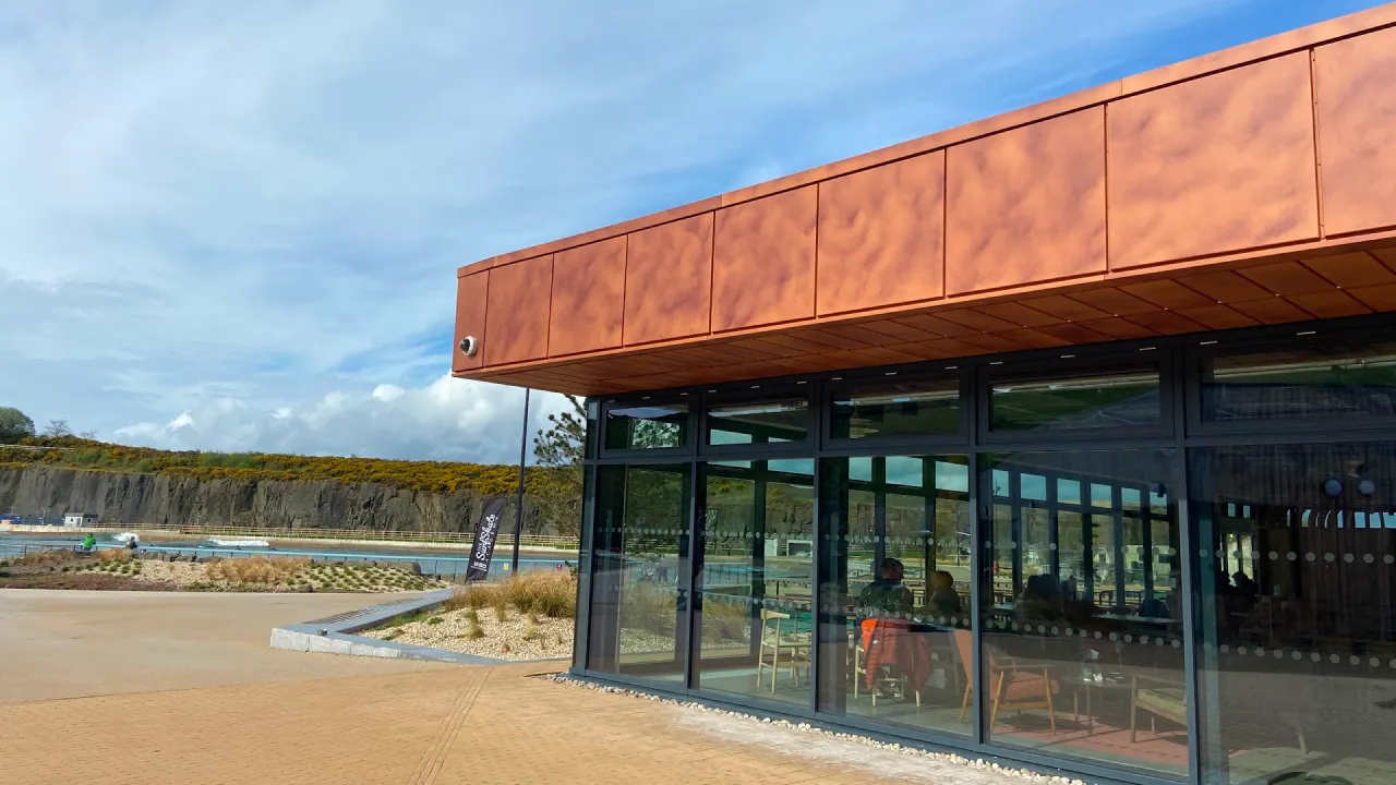 Modern glass-fronted café building at Lost Shore Surf Resort in Scotland, with copper-toned cladding, indoor seating visible through large windows, and the surf lagoon and rocky hillside in the background under a bright sky.