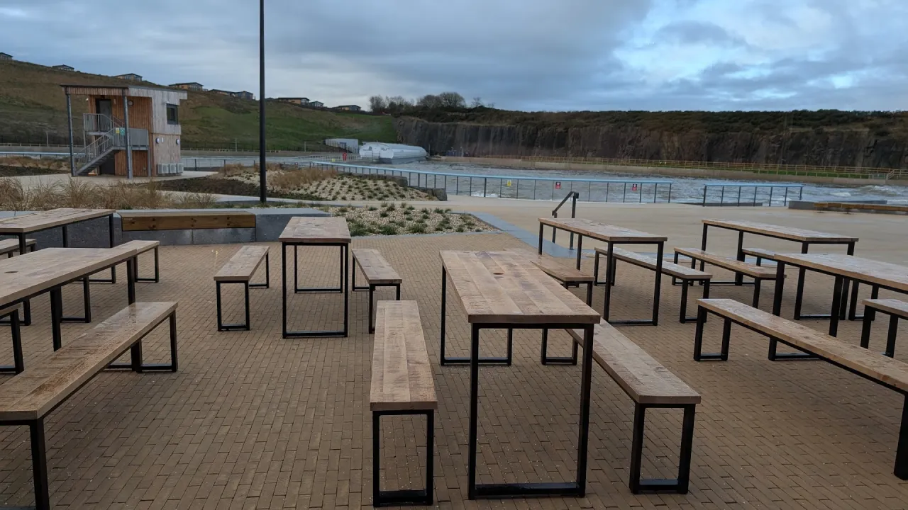 Wooden picnic tables and benches on the outdoor terrace at Lost Shore Surf Resort in Scotland, with the surf lagoon and waterfront walkway behind them, landscaped planting beds, and a rocky cliff line under a cloudy sky.