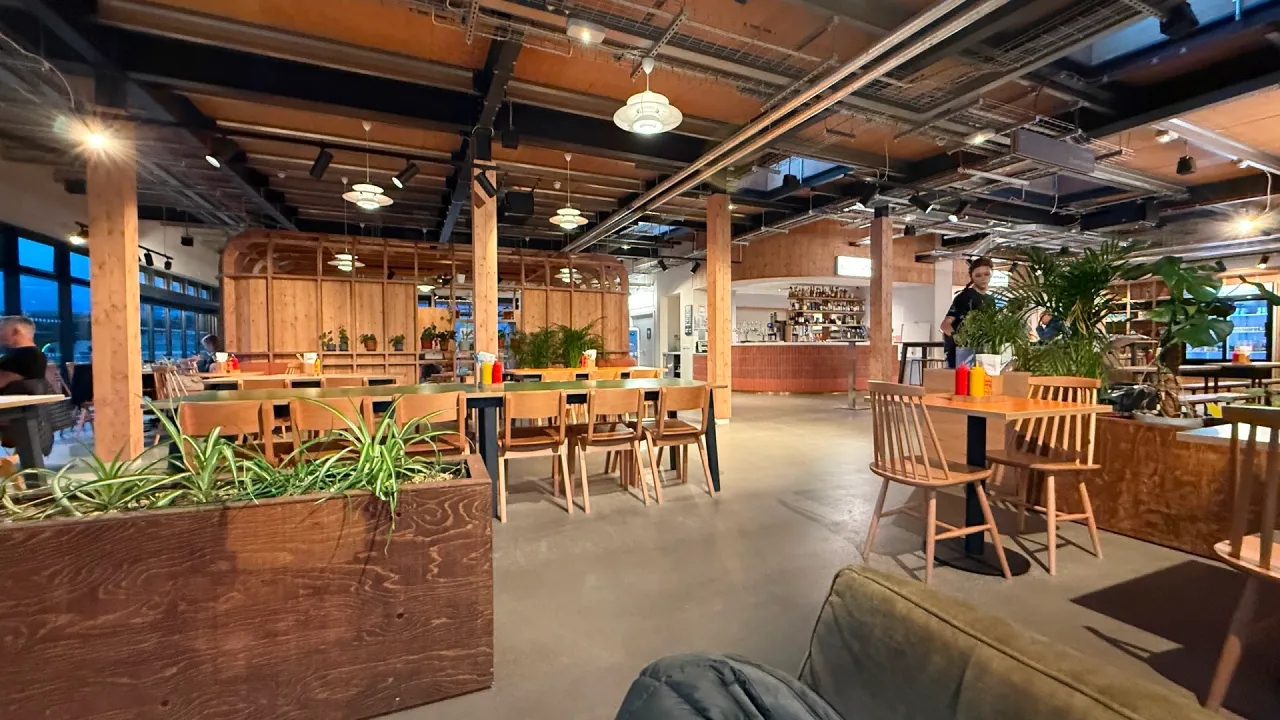 Open-plan café and bar interior at Lost Shore Surf Resort in Scotland, with wooden beams, communal tables and chairs, indoor planters, and a service counter under ceiling lights.