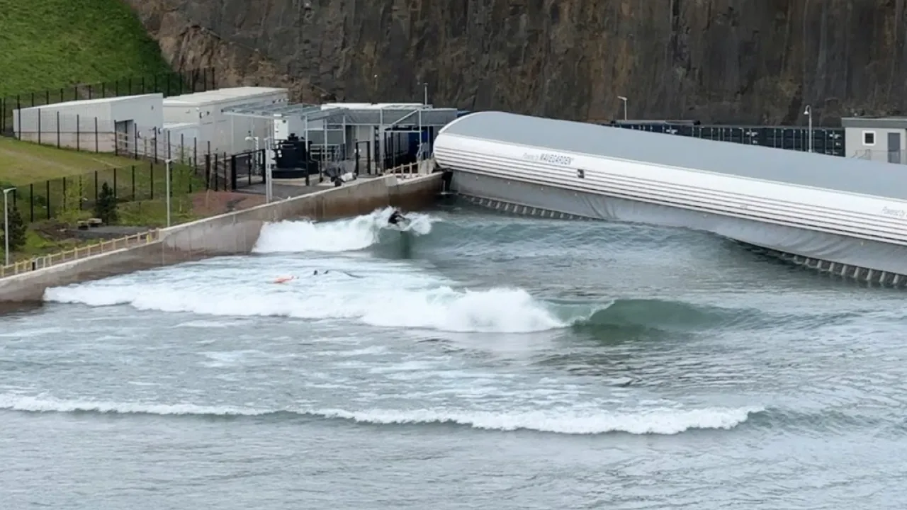 Surfer riding a breaking wave in the Lost Shore Surf Resort surf lagoon in Scotland, with the wave generator visible at the end of the pool.