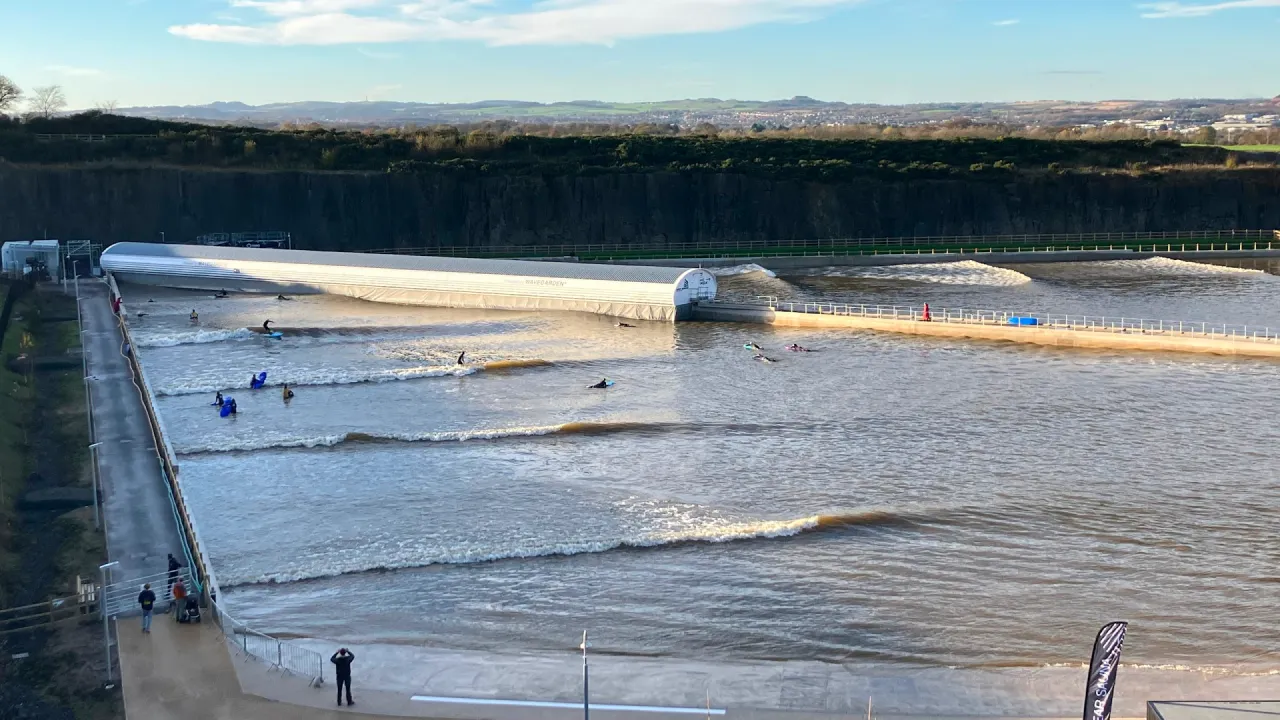 Panoramic view of the Lost Shore Surf Resort surf lagoon in Scotland, showing multiple surfers riding evenly spaced waves, with the wave generator at the far end.