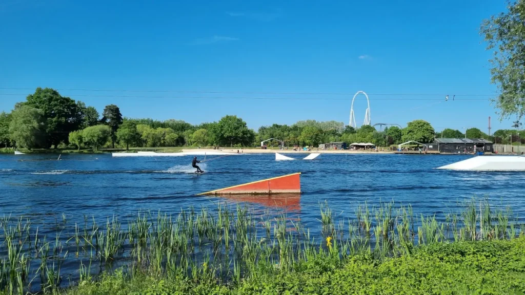 Wide lake view at Thorpe Lakes with a wakeboarder on the water, floating ramps and obstacles, shoreline greenery, and the tall rollercoaster structure of Thorpe Park in the distance.