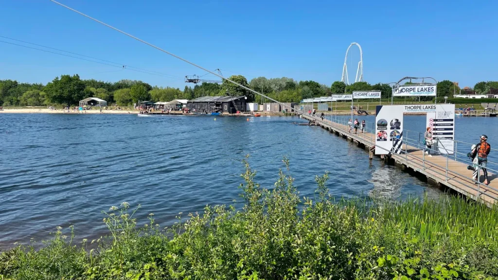 Wide view of Thorpe Lakes showing the water, a long boardwalk with Thorpe Lakes signs, and the watersports facilities and cable system in the background.