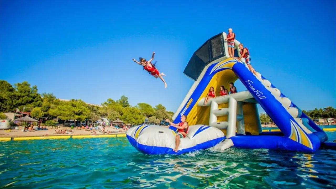 Person leaping from the ‘Freefall’ inflatable at Thorpe Lakes Aqua Park, with others in buoyancy aids on the structure, and the lakeside beach in the background.