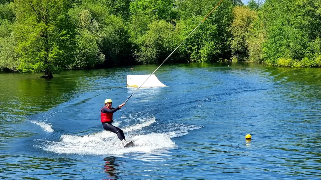 Wakeboarder in a red buoyancy aid being pulled by the cable system at Thorpe Lakes, with a floating ramp obstacle and tree-lined shore in the background.