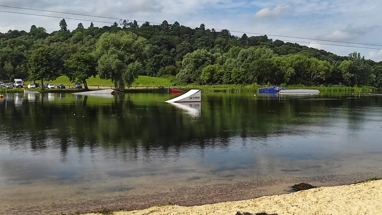 Wide shot of Thorpe Lakes with glassy water reflecting trees, sandy beach in the foreground, and several wake park obstacles and ramps positioned across the lake.