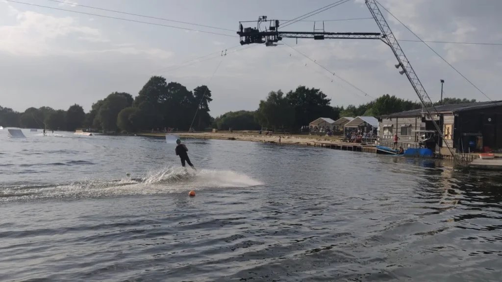 Wakeboarder being pulled across Thorpe Lakes by the overhead cable system, creating a spray trail, with the shoreline and watersports buildings in the background.