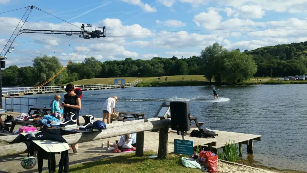 Visitors relaxing and preparing wakeboarding gear on the lakeside beach deck at Thorpe Lakes, with a cable wakeboarder riding across the lake behind them.