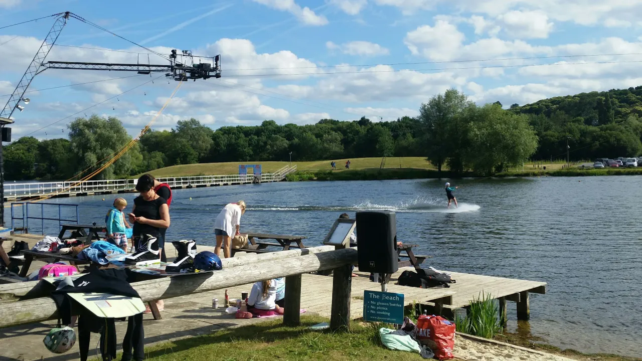 Visitors relaxing and preparing wakeboarding gear on the lakeside beach deck at Thorpe Lakes, with a cable wakeboarder riding across the lake behind them.